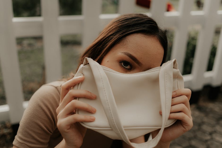 Close-up of a woman playfully hiding her face with a white handbag in an outdoor setting.